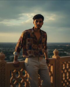 Create a retro vintage-style, grainy but bright photograph of me (use image for face)in a 90s shirt and a little casualfut pant. The setting should be a [dramatic background, e.g., grand palace balcony, lush garden]. The lighting should be dramatic and cinematic, with deep shadows and warm, golden highlights, reminiscent of a 90s Bollywood film still. Her expression should be [mood, e.g., mysterious and alluring, softly smiling]."